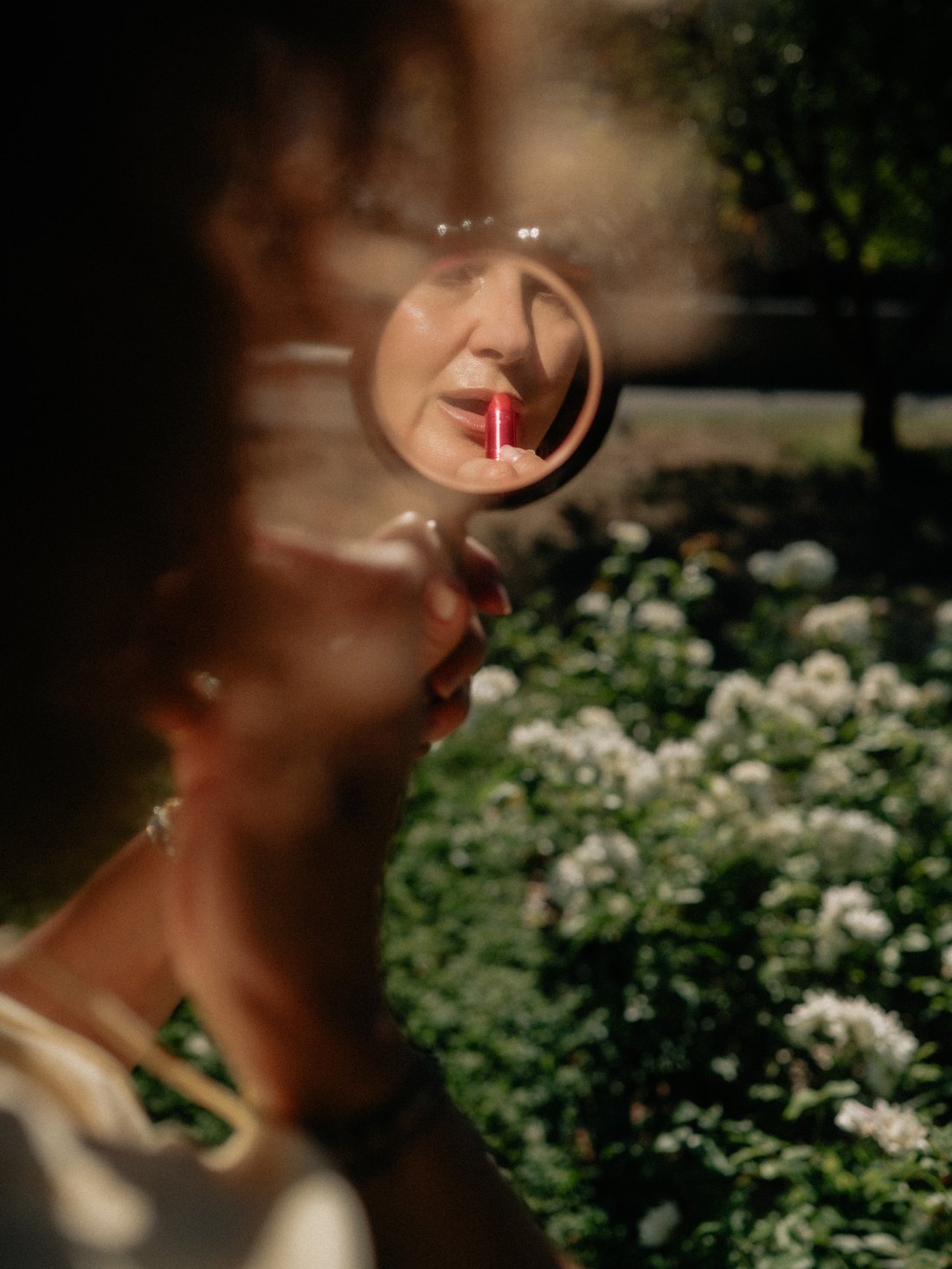 Une femme applique son rouge à lèvres dans un petit miroir rond, à contre-jour dans un jardin
